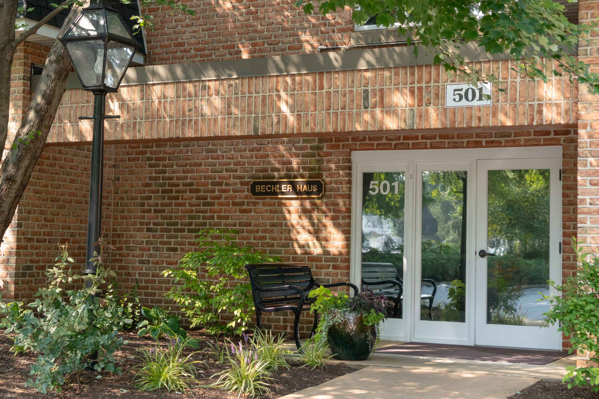 Entrance to a brick building with the number 501 displayed above and on the glass doors. There is a black bench and a large planter with greenery near the entrance. A black lamp post and various plants and shrubs are visible in front of the building.