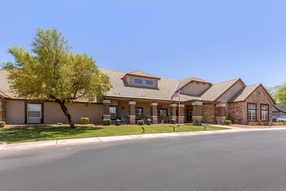 Exterior front view of a single-story senior living facility building with a stone and stucco facade, a sloped roof, a small porch area with columns, an American flag on a flagpole, and a tree on a well-maintained lawn under a clear blue sky.
