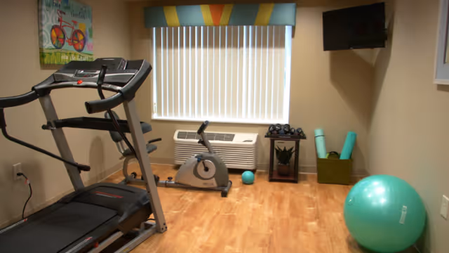Small exercise room with a treadmill, stationary bike, set of dumbbells on a stand, exercise balls, yoga mats in a basket, a wall-mounted TV, and a window with vertical blinds and a colorful valance.