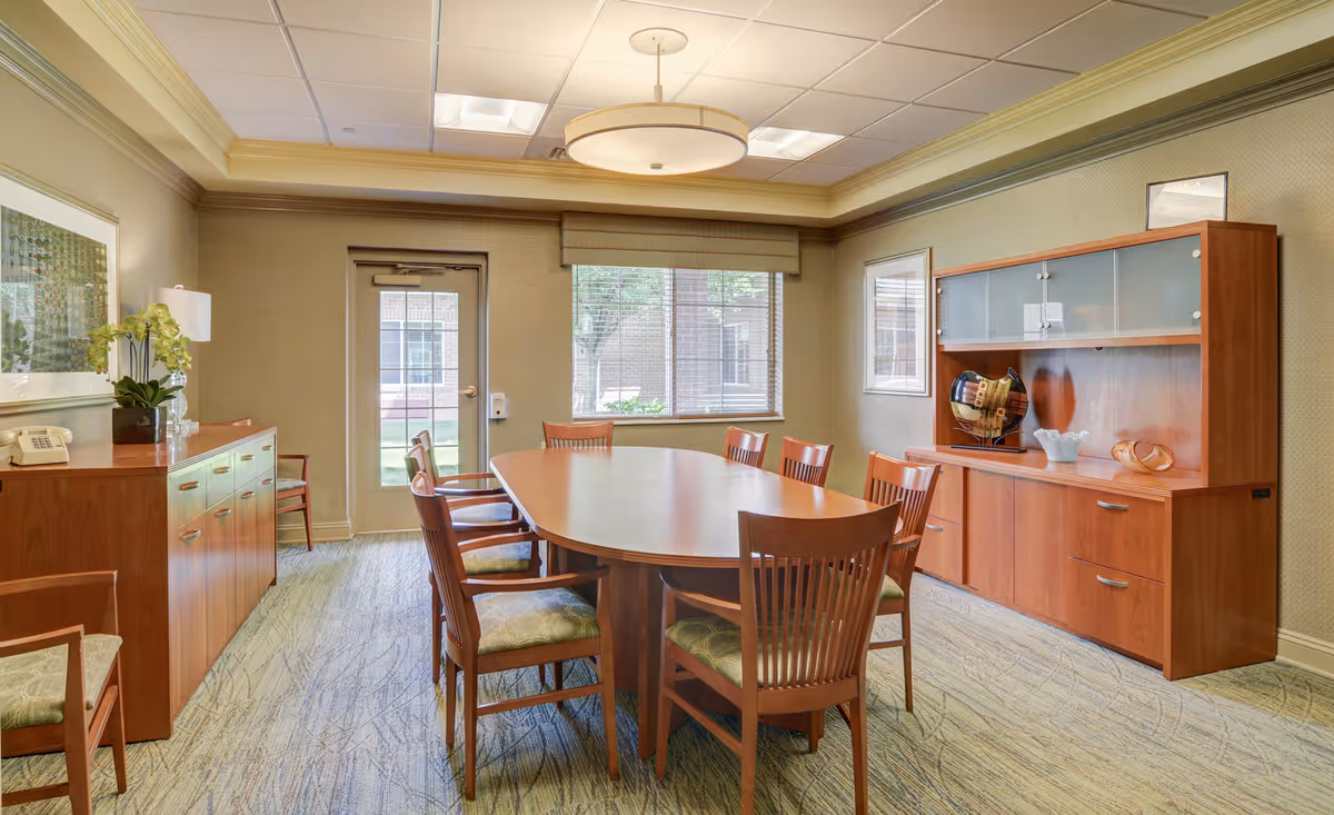 A well-lit meeting or dining room with a large wooden oval table surrounded by eight wooden chairs with green cushions. The room has beige walls, a patterned carpet, a wooden sideboard with a lamp and plant, and a wooden cabinet with decorative items. There is a door and a window showing an outdoor view.
