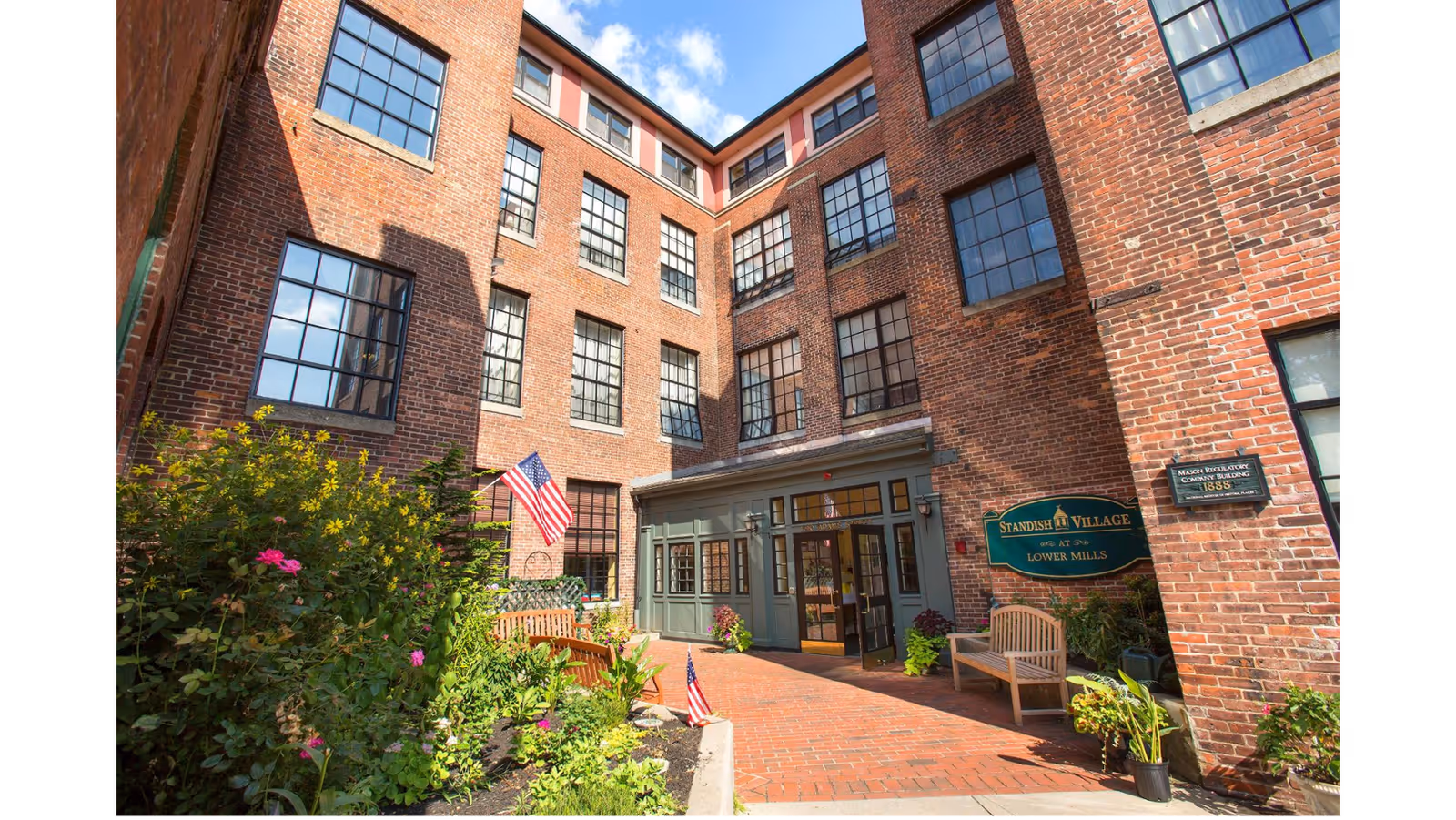 Exterior view of a brick building with multiple large windows and a small garden area with flowers and plants. There are two wooden benches near the entrance, which has a green sign reading 'Standish Village at Lower Mills.' An American flag is displayed near the garden. The sky is blue with some clouds.