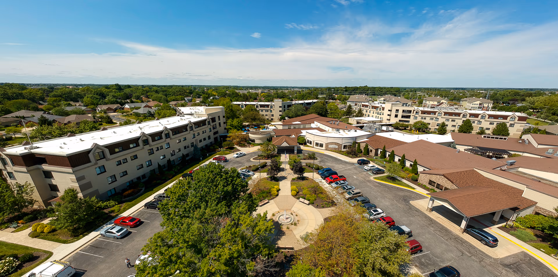 Aerial view of Westminster Village senior living facility showing multiple connected buildings with white and brown roofs, surrounded by trees and parking lots with cars. The landscape includes green lawns and a circular courtyard with a fountain in the center.