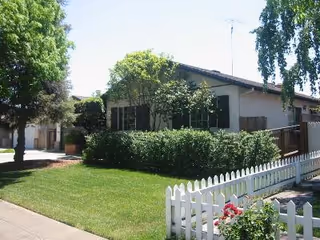 Single-story residential building with a white picket fence, green lawn, and trees surrounding the property under a clear sky.