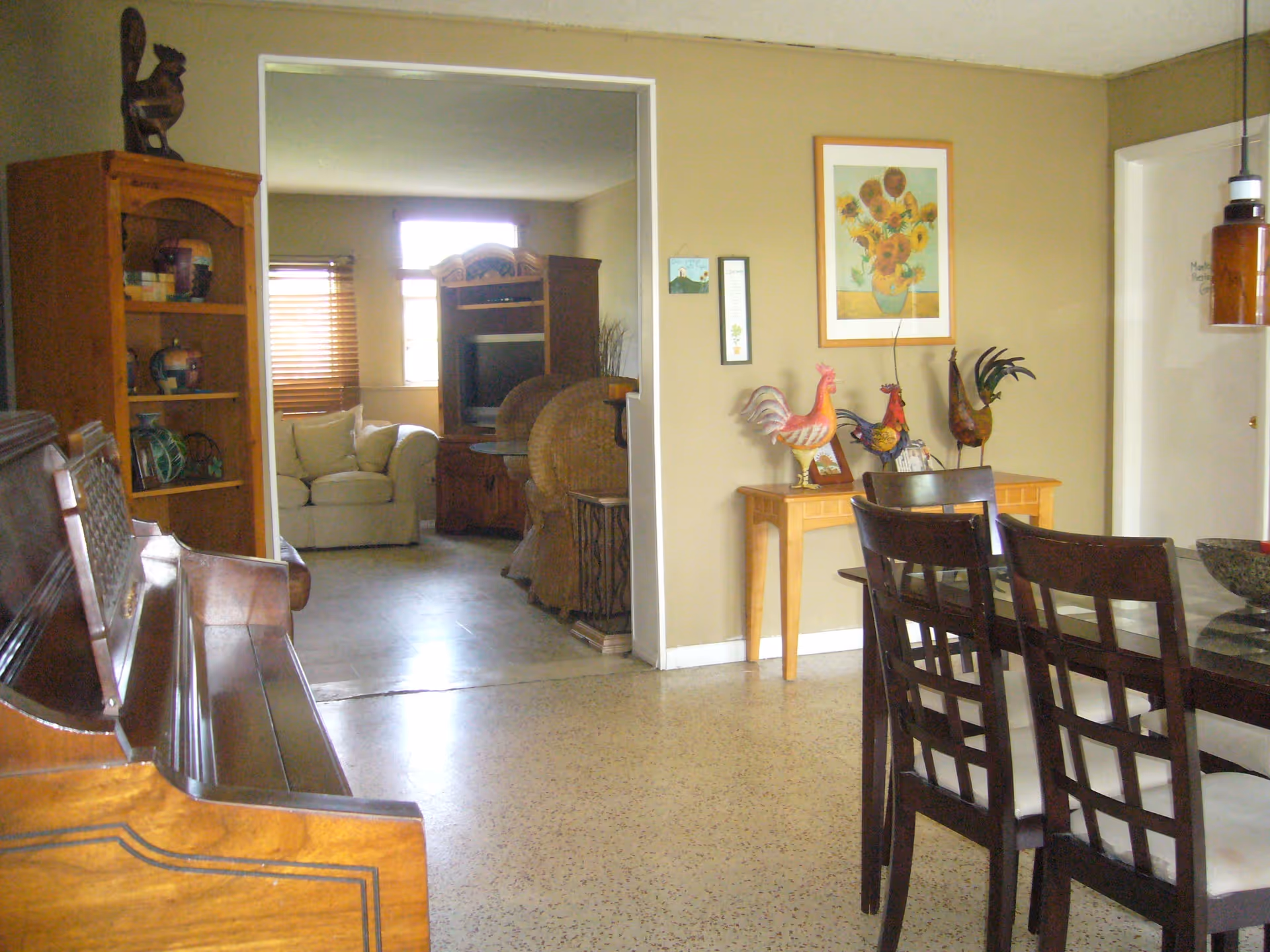 Interior view of a living and dining area in a senior living facility. The foreground shows a dining table with dark wooden chairs and a glass top. To the left, there is a wooden piano and a wooden bookshelf with decorative items. In the background, a cozy living room is visible with a beige armchair, a wooden entertainment center, and wicker chairs. The walls are beige with framed artwork and decorative rooster figurines on a small wooden table.