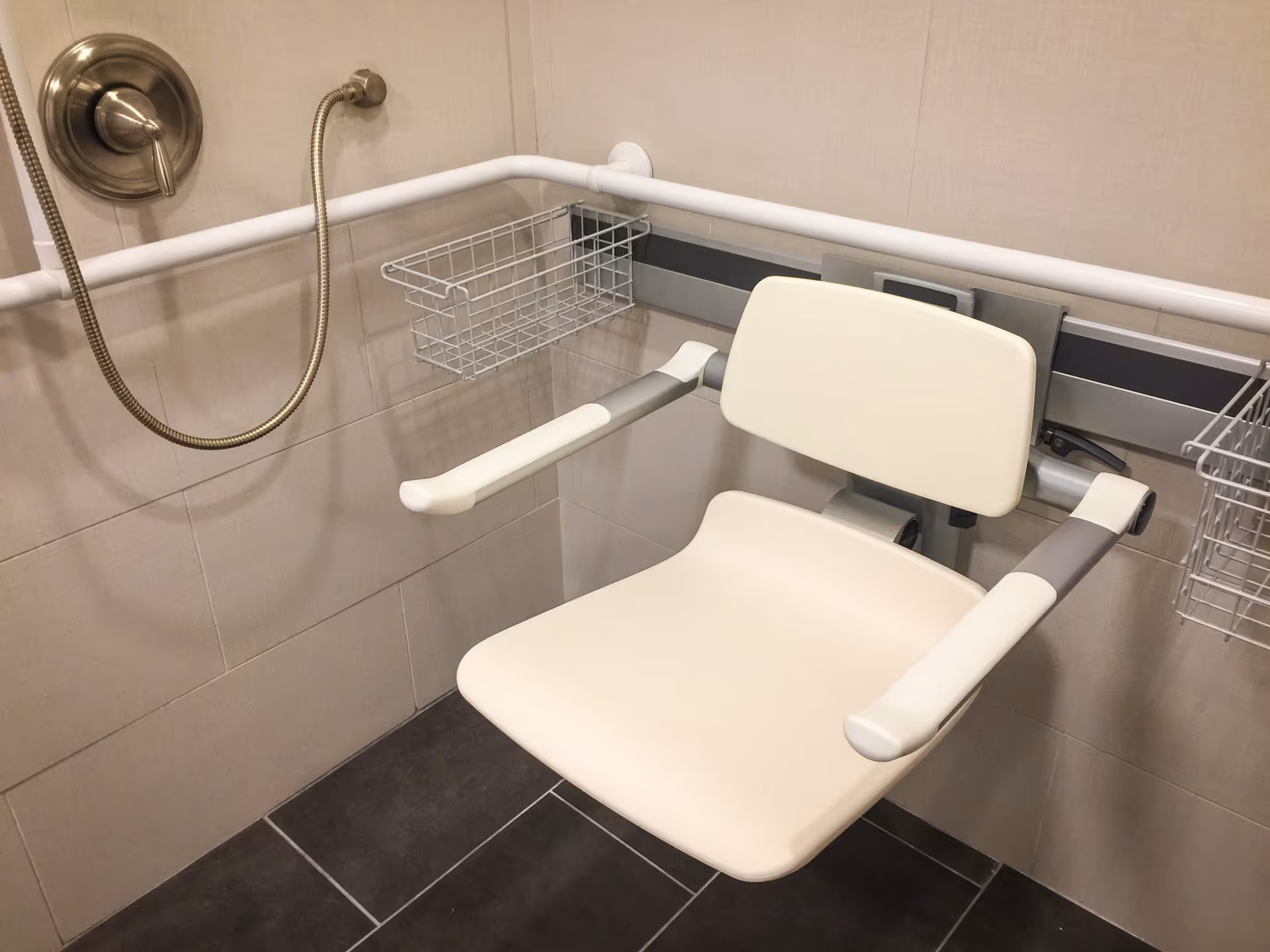 A beige and gray foldable shower seat mounted on a tiled bathroom wall with white grab bars and a handheld showerhead with a metal hose. There are two wire baskets attached to the wall for holding shower items.