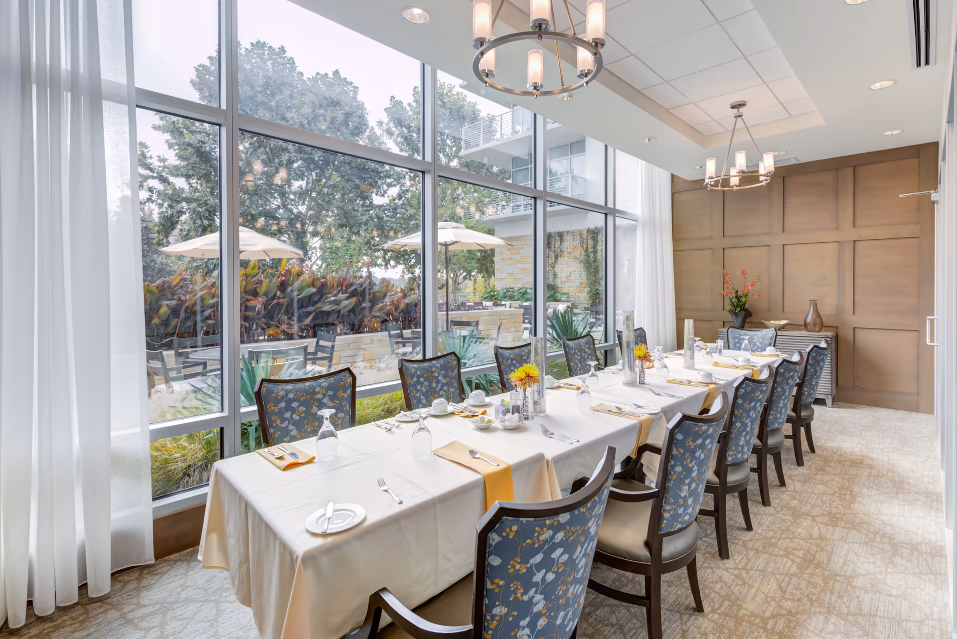 A bright dining room with a long table set for a meal, featuring blue floral upholstered chairs, white tablecloth, yellow napkins, and glassware. Large floor-to-ceiling windows provide a view of an outdoor patio with umbrellas, plants, and trees. The room has modern chandeliers and a wooden paneled accent wall with decorative vases and flowers.