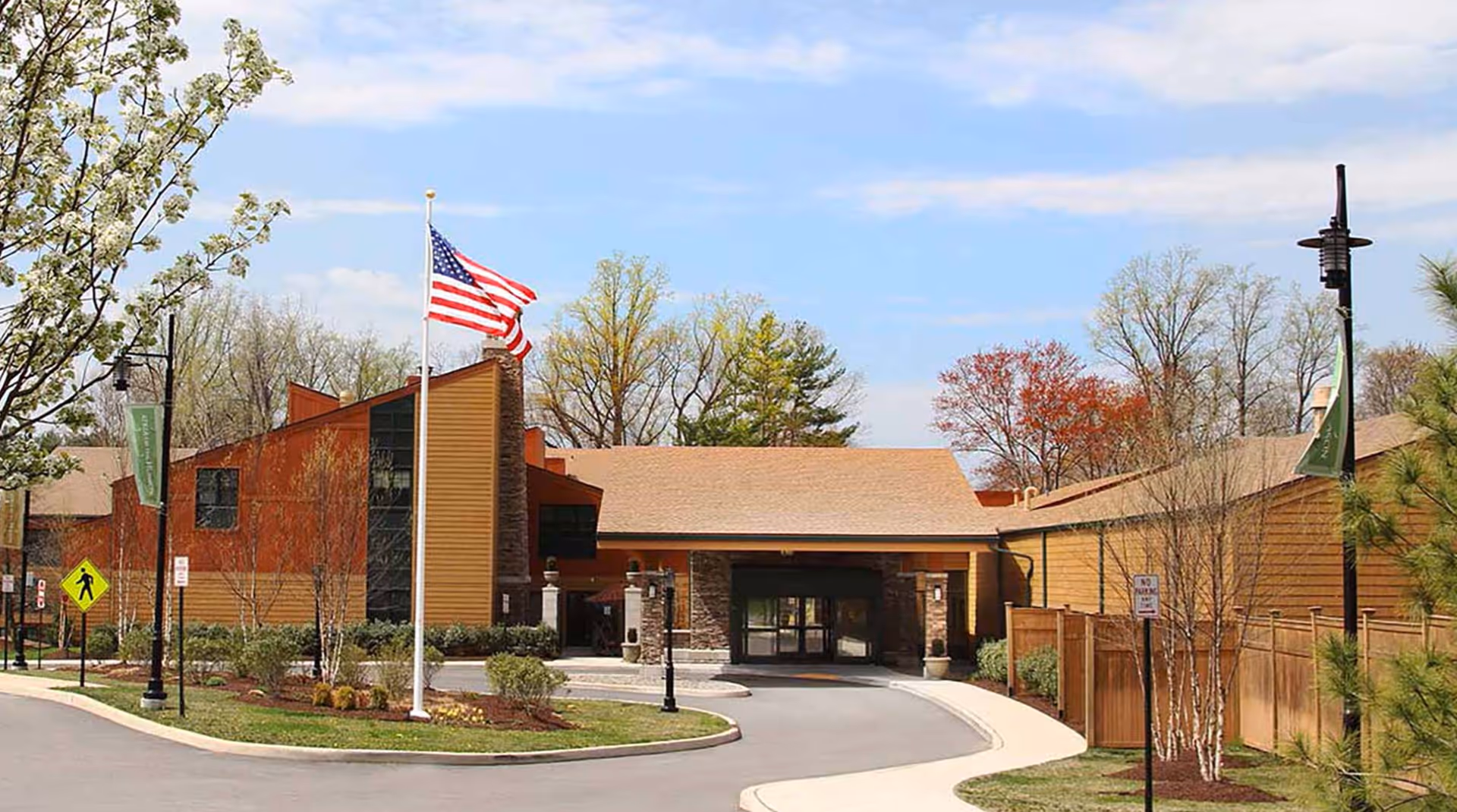 Exterior view of a senior living facility building with a driveway, landscaped greenery, an American flag on a flagpole, and trees in the background under a partly cloudy sky.