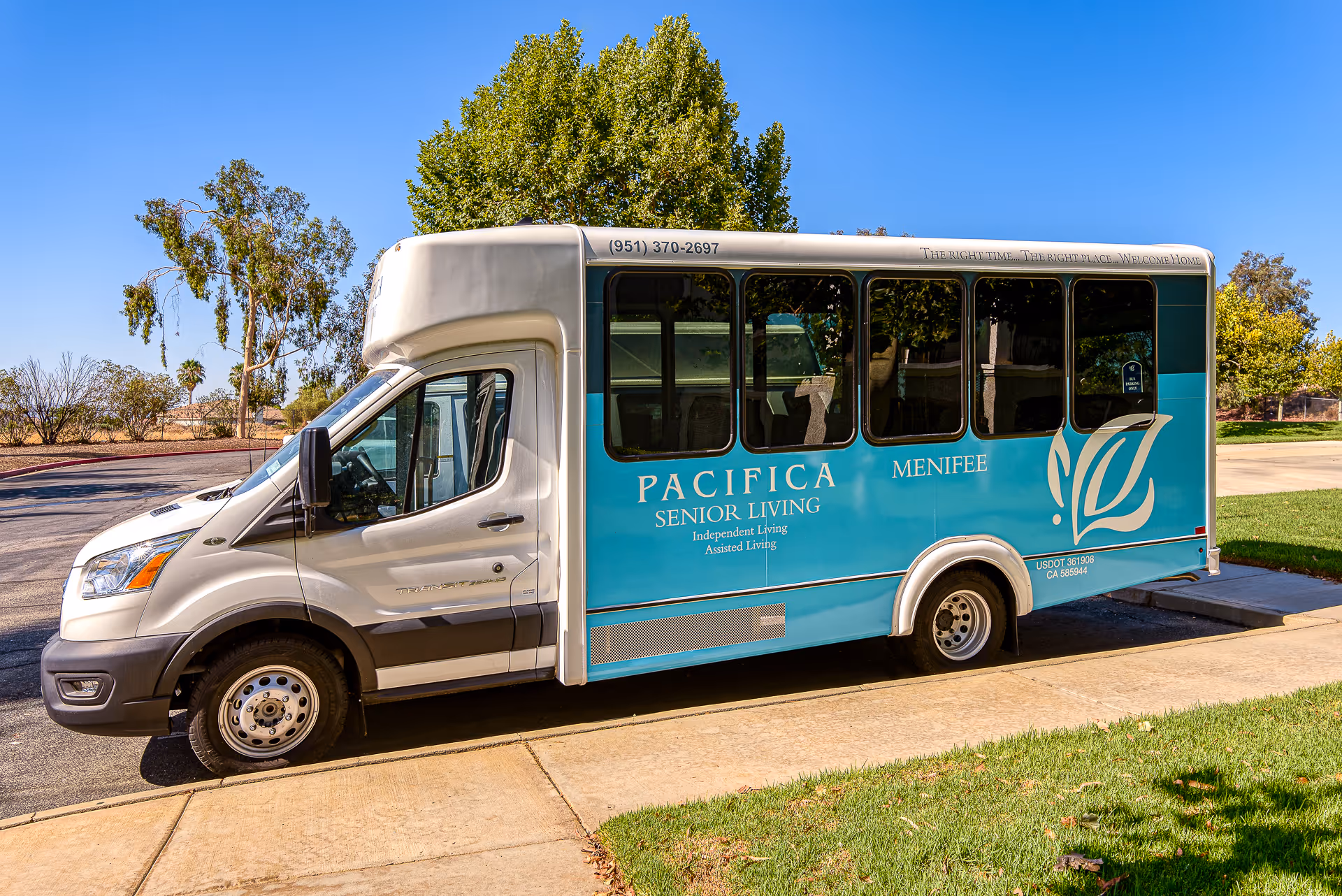 A white and teal shuttle bus parked on a street next to a sidewalk and grass. The bus has the text 'Pacifica Senior Living Menifee' along with 'Independent Living Assisted Living' on its side. Trees and a clear blue sky are visible in the background.