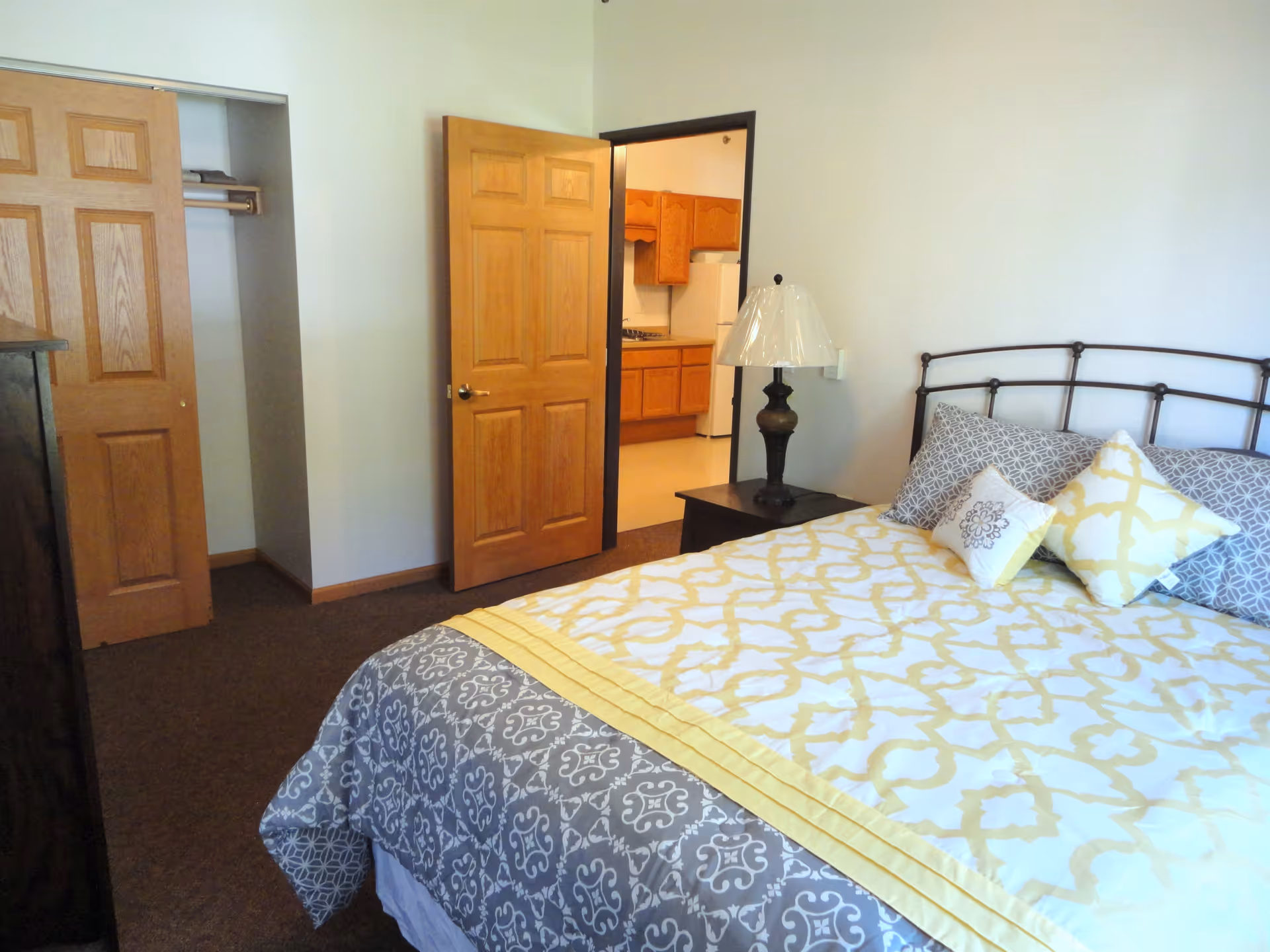 A bedroom with a bed covered in a yellow and gray patterned comforter and several pillows. There is a wooden nightstand with a lamp next to the bed. The room has a wooden door that is open, revealing a kitchen area with wooden cabinets and a white refrigerator. There is also an open closet with sliding wooden doors.