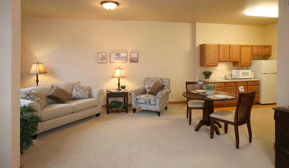 A cozy living area with a beige sofa adorned with patterned and solid pillows, a matching armchair, and two table lamps on side tables. The room features a small round dining table set with two chairs and plates, adjacent to a compact kitchen with wooden cabinets, a microwave, and a refrigerator. The walls are light-colored, and the floor is carpeted.