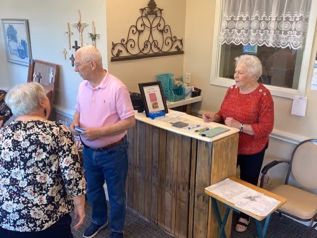 Three elderly people interacting in a cozy room with a wooden reception desk. One woman in a red sweater stands behind the desk, while a man in a pink polo shirt and a woman in a floral top stand in front of it. The room has decorative wall art, a window with lace curtains, and a small table with papers and pens.