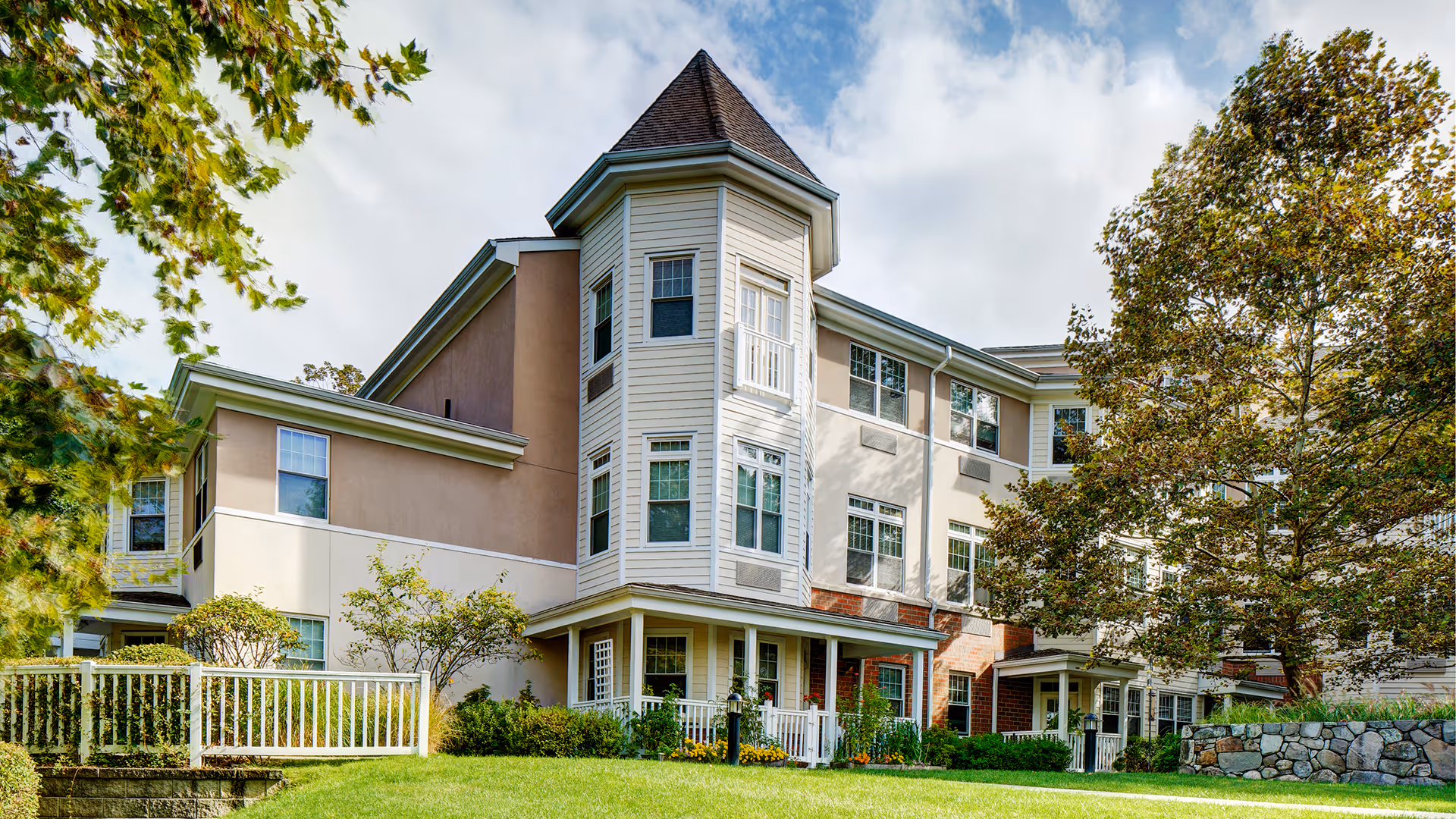 Exterior view of a multi-story senior living facility building with beige and light brown walls, multiple windows, a turret-like architectural feature, surrounded by green trees and a well-maintained lawn with a white fence.