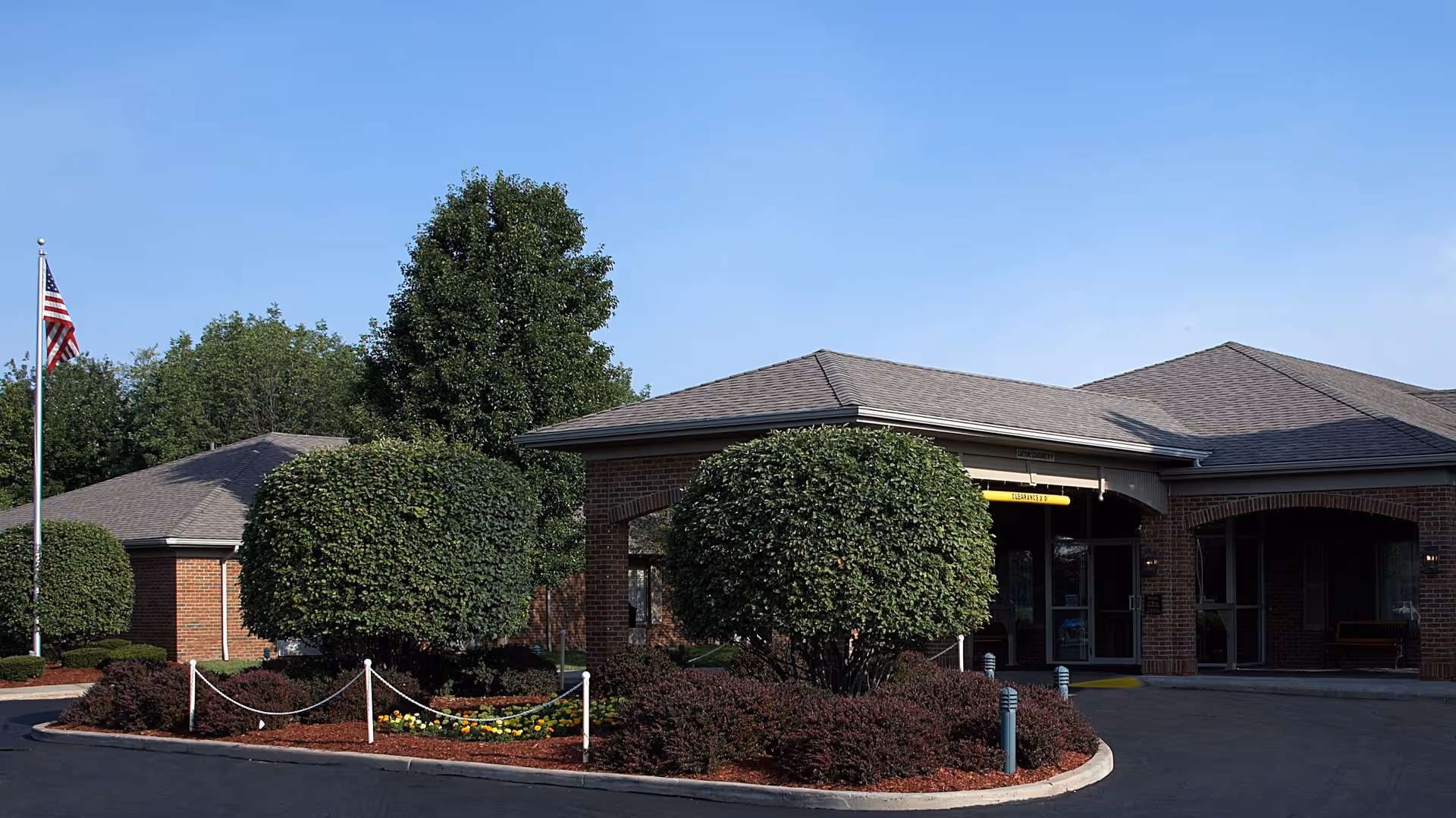 Exterior view of a senior living facility building with a circular driveway, neatly trimmed bushes, a flagpole with an American flag, and a clear blue sky.