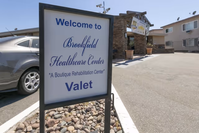 A sign at the entrance of Brookfield Healthcare Center, a boutique rehabilitation center, indicating valet parking. The sign is placed on a bed of rocks next to a parking lot with a silver car and the facility building in the background under a clear blue sky.