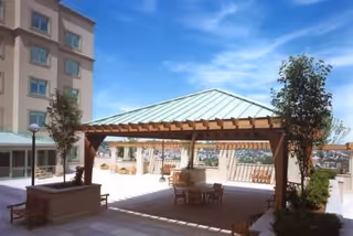 Outdoor patio area with a wooden pergola providing shade over a table and chairs, surrounded by potted plants and trees, with a multi-story building in the background under a clear blue sky.