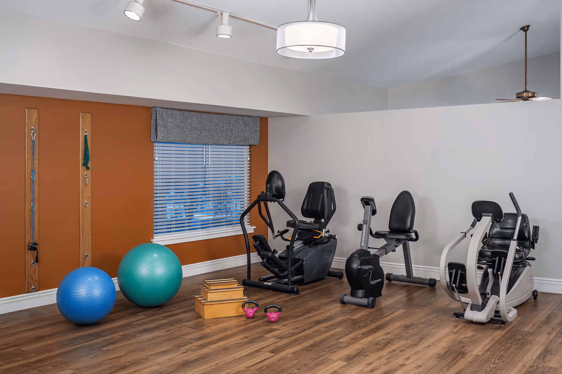A senior living facility exercise room with wooden flooring, featuring two large exercise balls, resistance bands mounted on the wall, two kettlebells, wooden step platforms, and three pieces of exercise equipment including a recumbent bike and a rowing machine. The room has a window with blinds and a gray valance, white and orange walls, and ceiling lights.