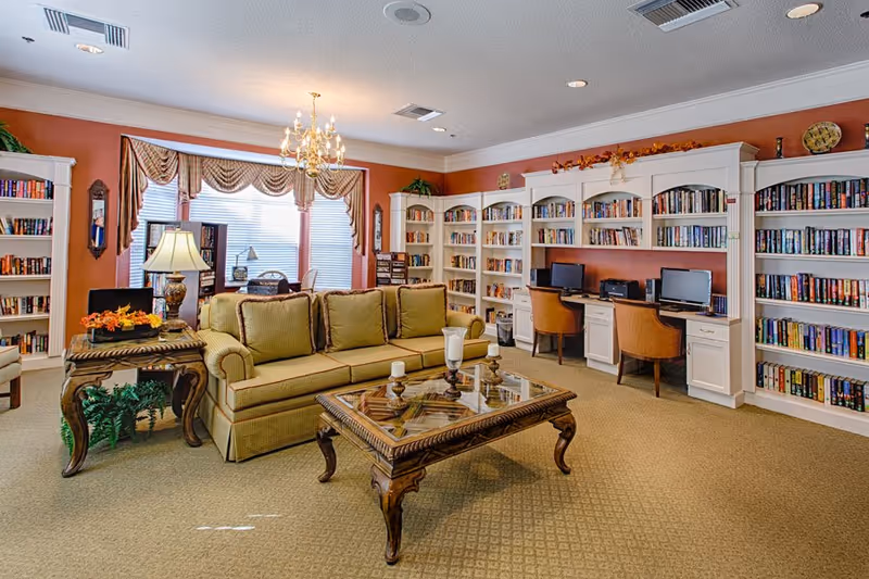 A cozy senior living facility common area with a green sofa, a wooden coffee table with candles, and a side table with a lamp and flowers. The room features built-in white bookshelves filled with books and two computer workstations with chairs. Large windows with draped curtains allow natural light to fill the space.