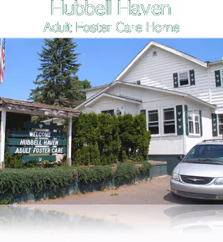 Exterior view of Hubbell Haven Adult Foster Care Home showing a white building with green shutters, a sign that reads 'Welcome Hubbell Haven Adult Foster Care', some greenery, and a parked silver car.