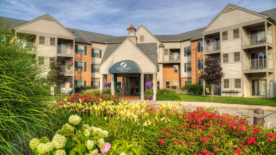 Front exterior view of Oakmont Manor Senior Community building with a covered entrance, surrounded by well-maintained gardens featuring various colorful flowers and greenery under a partly cloudy sky.