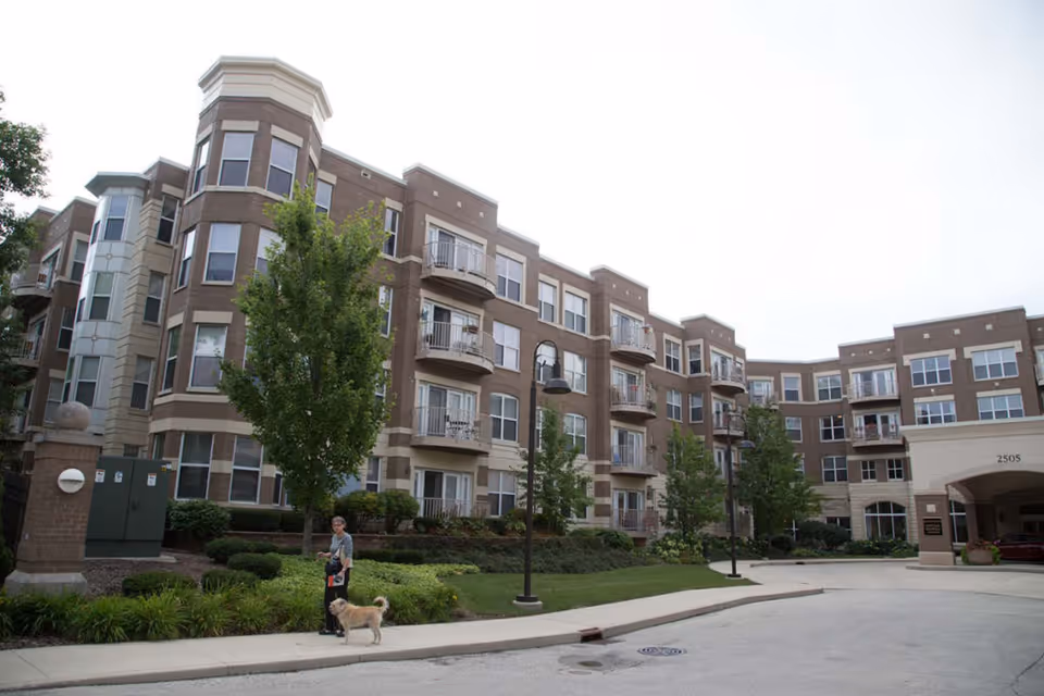 Exterior view of a multi-story senior living facility named Eastcastle Place with balconies, trees, and landscaped greenery. A person is walking a dog on the sidewalk in front of the building.