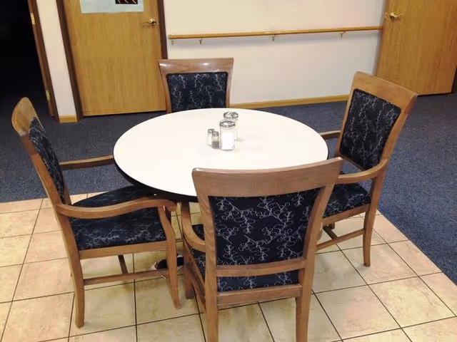A round white table with four wooden chairs featuring dark blue patterned cushions arranged around it. The table has salt and pepper shakers and a sugar dispenser on top. The room has tiled flooring near the table and carpeted flooring further back, with two wooden doors and a handrail on the wall in the background.