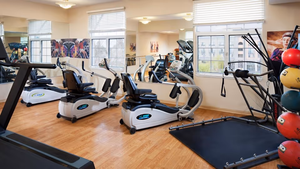 A bright fitness room with wooden flooring featuring exercise equipment including two NuStep recumbent cross trainers, a treadmill, a multi-functional workout machine, and a rack of colorful medicine balls. Large windows and mirrors line the walls, allowing natural light to fill the space.