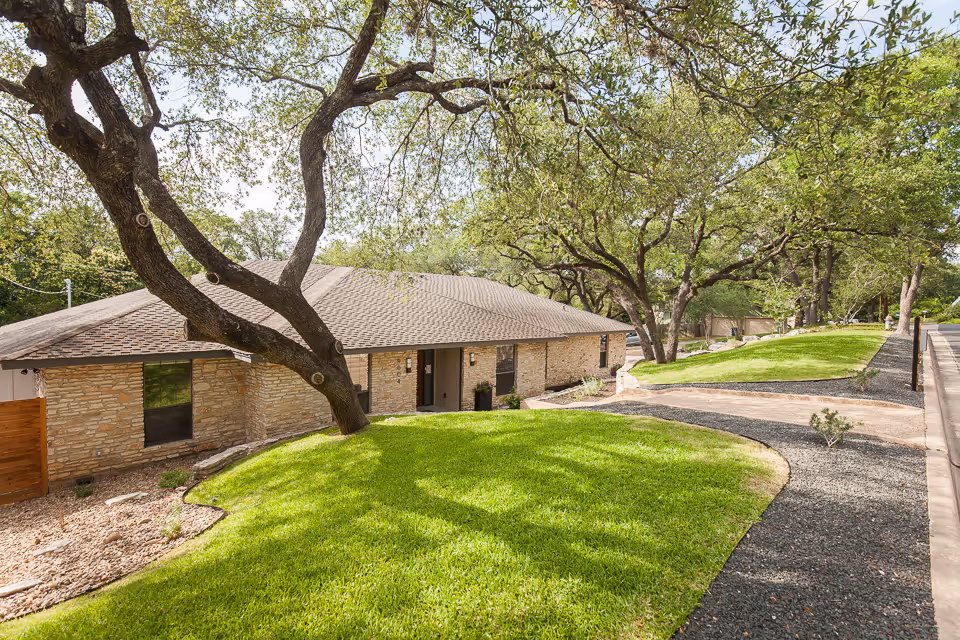 Exterior view of a single-story stone building with a shingled roof surrounded by well-maintained green lawns and large trees providing shade. A paved walkway curves along the front of the building, and the scene is bright and sunny.