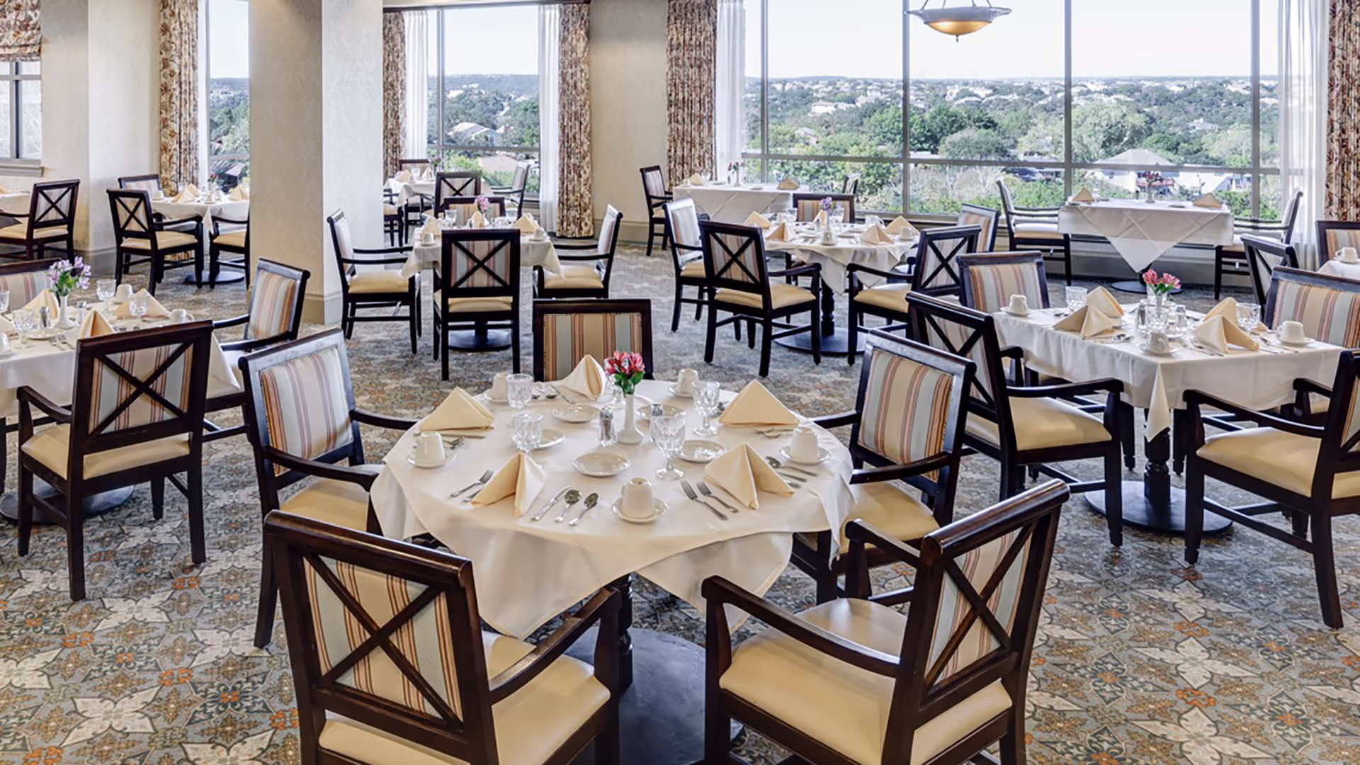 A bright dining room with multiple round and square tables set with white tablecloths, beige napkins, glassware, and silverware. The chairs have wooden frames with cushioned seats and striped backs. Large windows provide a scenic view of trees and distant buildings outside.