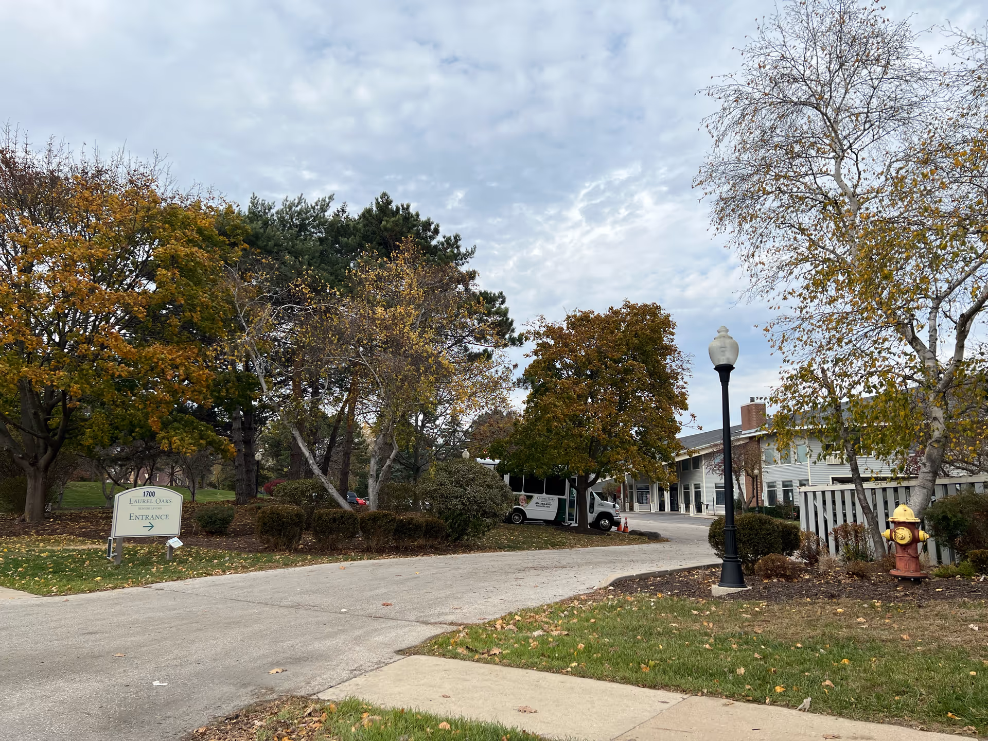 Exterior view of Laurel Oaks Senior Living facility showing a driveway entrance with a sign that reads '1700 Laurel Oaks Senior Living Entrance' surrounded by trees with autumn foliage, a street lamp, a fire hydrant, and a shuttle van parked near the building.