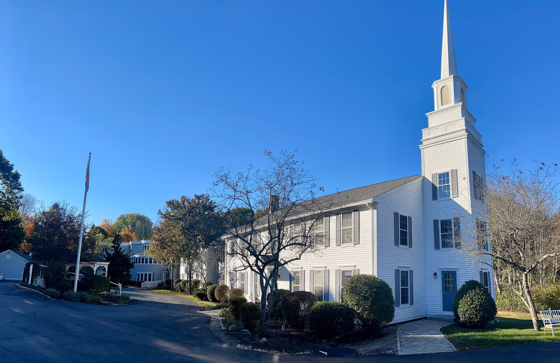 Exterior view of a white building with a tall steeple, surrounded by trees and bushes under a clear blue sky. There is a paved driveway and a flagpole in front of the building.