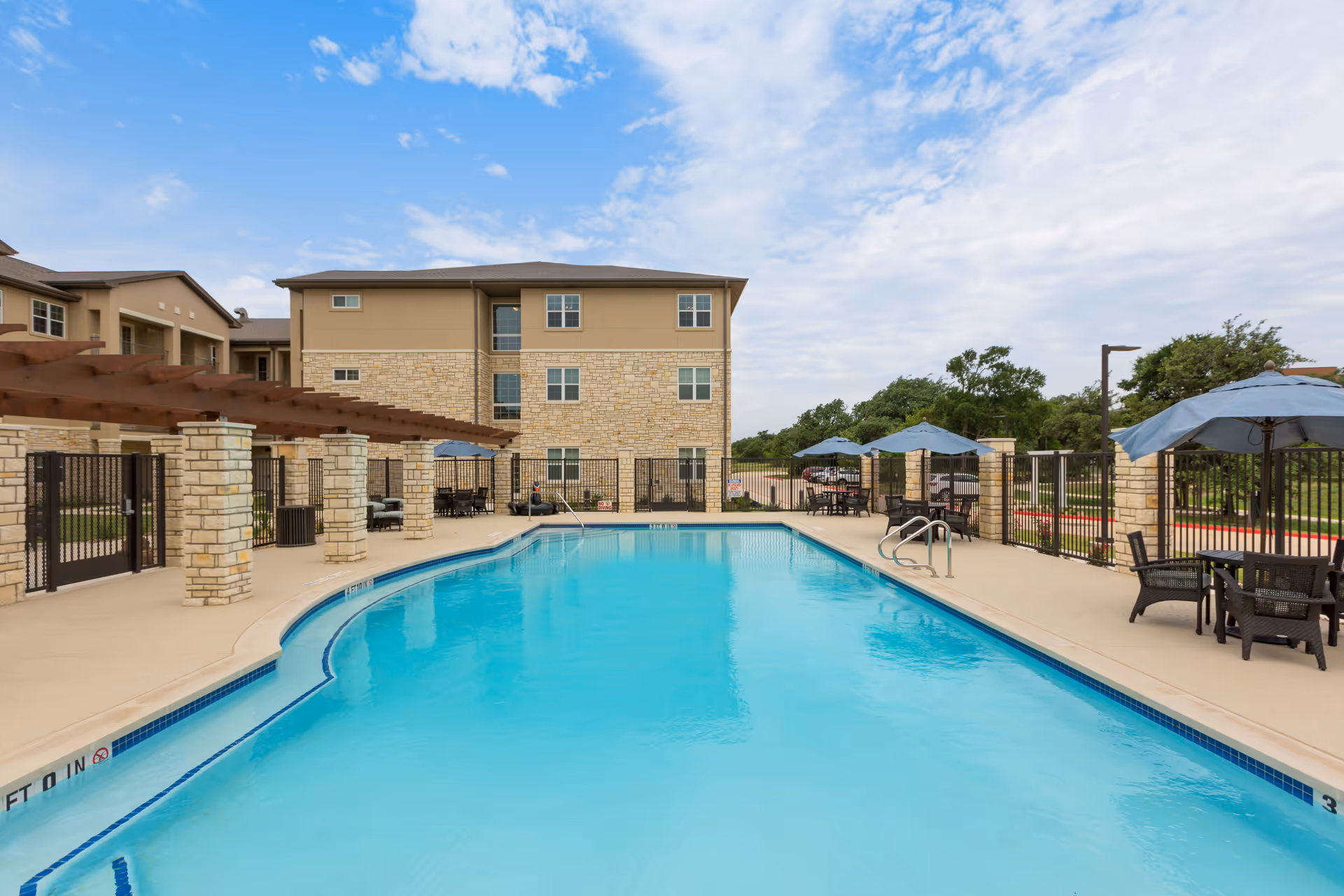 Outdoor swimming pool area at The Enclave at Round Rock Senior Living with clear blue water, surrounded by a concrete deck, patio tables with blue umbrellas, and a building with stone and beige exterior in the background under a partly cloudy sky.