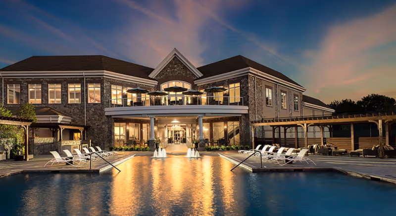 A large, elegant building with stone and brick exterior is shown at dusk with lights glowing warmly from inside. In front of the building is a swimming pool with water fountains and lounge chairs arranged on both sides. There are shaded seating areas with umbrellas on the balcony and pergolas on the pool deck. The sky is partly cloudy with a soft sunset glow.