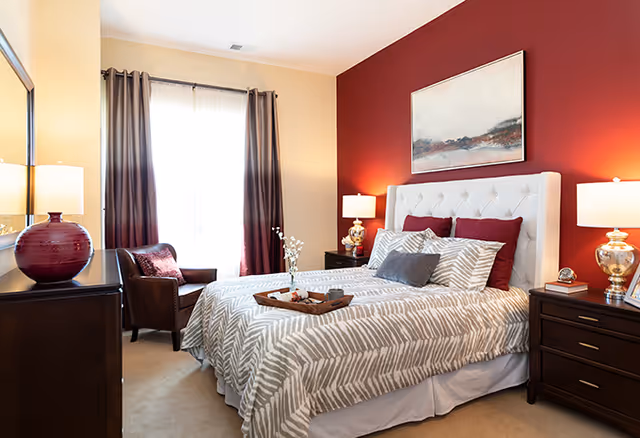 A cozy bedroom featuring a queen-sized bed with a tufted headboard, decorative pillows, and a striped comforter. The room has a warm color scheme with a red accent wall, two bedside lamps, and a wooden dresser. A comfortable armchair is positioned near a window with sheer curtains, adding to the inviting atmosphere.
