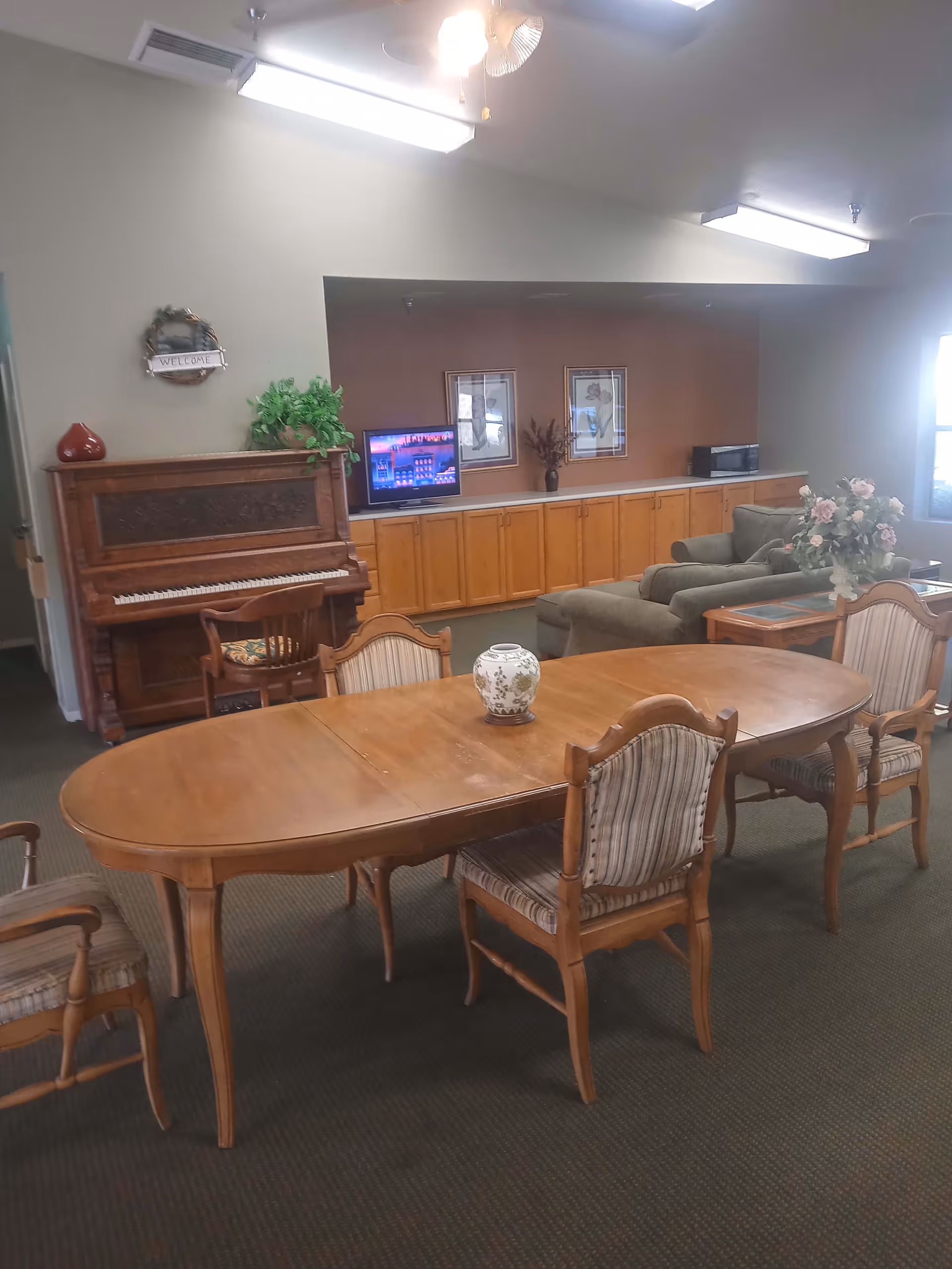A senior living facility common area featuring a wooden dining table with six chairs, a decorative vase on the table, a vintage upright piano with a chair, a small television on a long wooden cabinet, two framed pictures on the wall, a gray couch with a side table holding a flower arrangement, and a 'Welcome' wreath hanging on the wall.