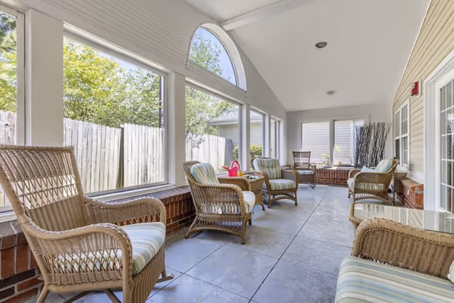 A bright enclosed patio area with large windows, featuring several wicker chairs with striped cushions arranged along the walls. The floor is tiled, and there is a view of a wooden fence and greenery outside the windows.