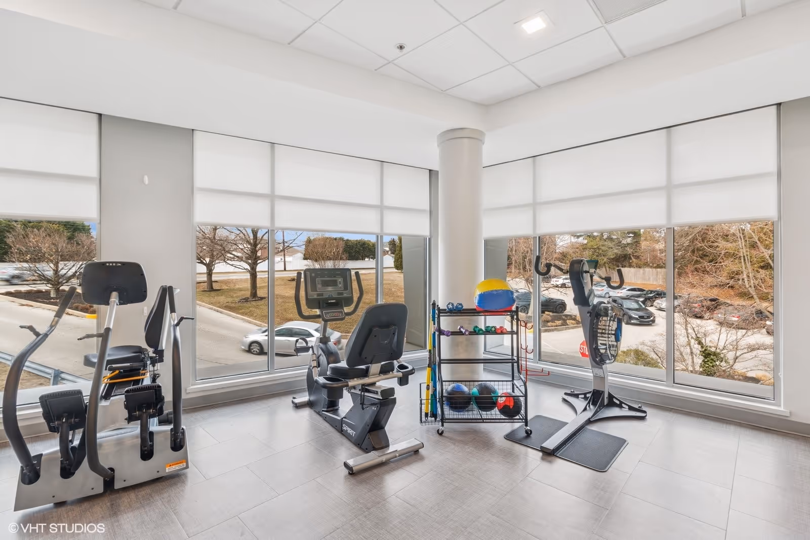 A bright exercise room with large windows showing an outdoor parking area. The room contains various fitness equipment including a recumbent bike, an elliptical machine, a stationary bike, and a rack holding colorful exercise balls and dumbbells.