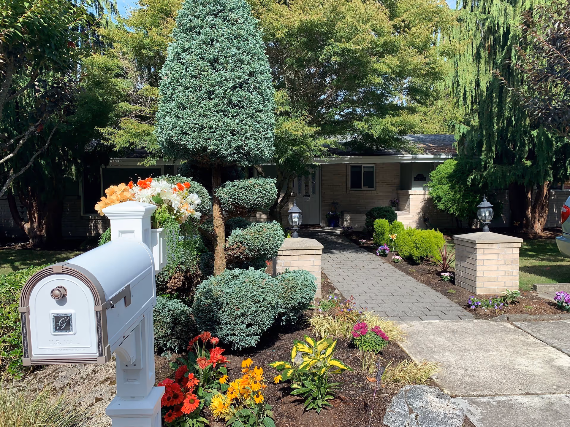 Front view of a single-story house with a well-maintained garden featuring colorful flowers and neatly trimmed bushes. A white mailbox with flowers on top is visible in the foreground, and a paved walkway leads to the front door flanked by two brick pillars with lanterns.
