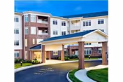 Exterior view of a multi-story senior living facility building with a covered entrance supported by brick columns, well-maintained landscaping, and a clear blue sky.
