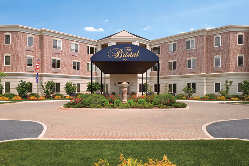 Front exterior view of The Bristal Assisted Living at Armonk building with a circular driveway, landscaped garden in the center, and a blue canopy with the facility's name and number 90.