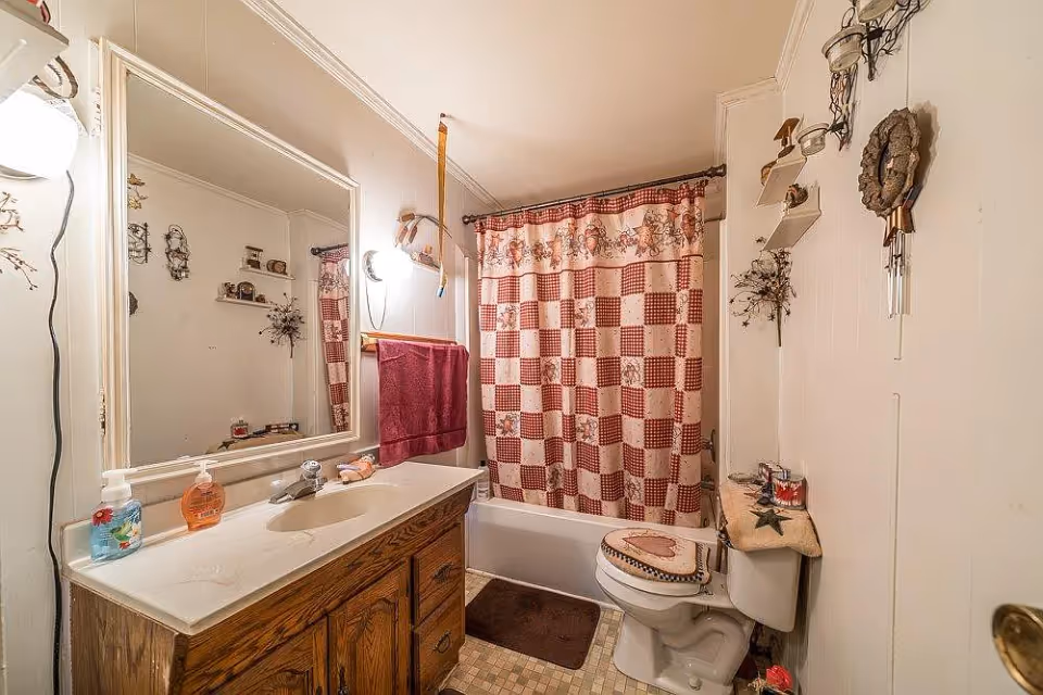 A bathroom with a wooden vanity and white countertop holding two soap dispensers. Above the vanity is a large mirror with a light fixture on the left side. A red towel hangs on a rack next to the mirror. The bathtub has a red and white checkered shower curtain with a floral pattern. The toilet has a decorative cover with a heart design, and there are shelves on the wall holding small decorative items. The floor has small square tiles in beige tones.
