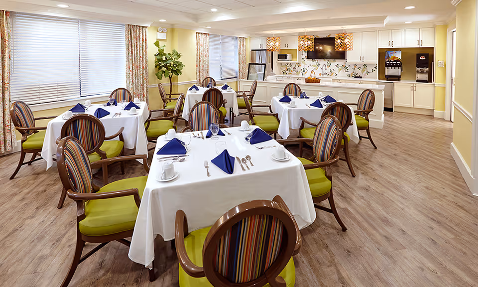 A bright and clean dining room with several tables covered in white tablecloths, each set with blue napkins, cups, and silverware. The chairs have wooden frames with striped upholstery and green cushions. The room features large windows with floral curtains, a wooden floor, and a kitchenette area with white cabinets, a microwave, a TV, and a beverage dispenser.
