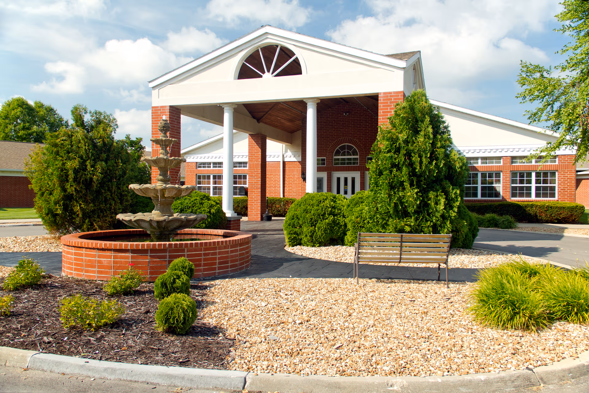 Front exterior view of Wilshire At Lakewood Rehab Center showing a brick building with white columns supporting a covered entrance. There is a multi-tiered water fountain surrounded by landscaping with bushes, small trees, and gravel. A bench is placed near the entrance area.