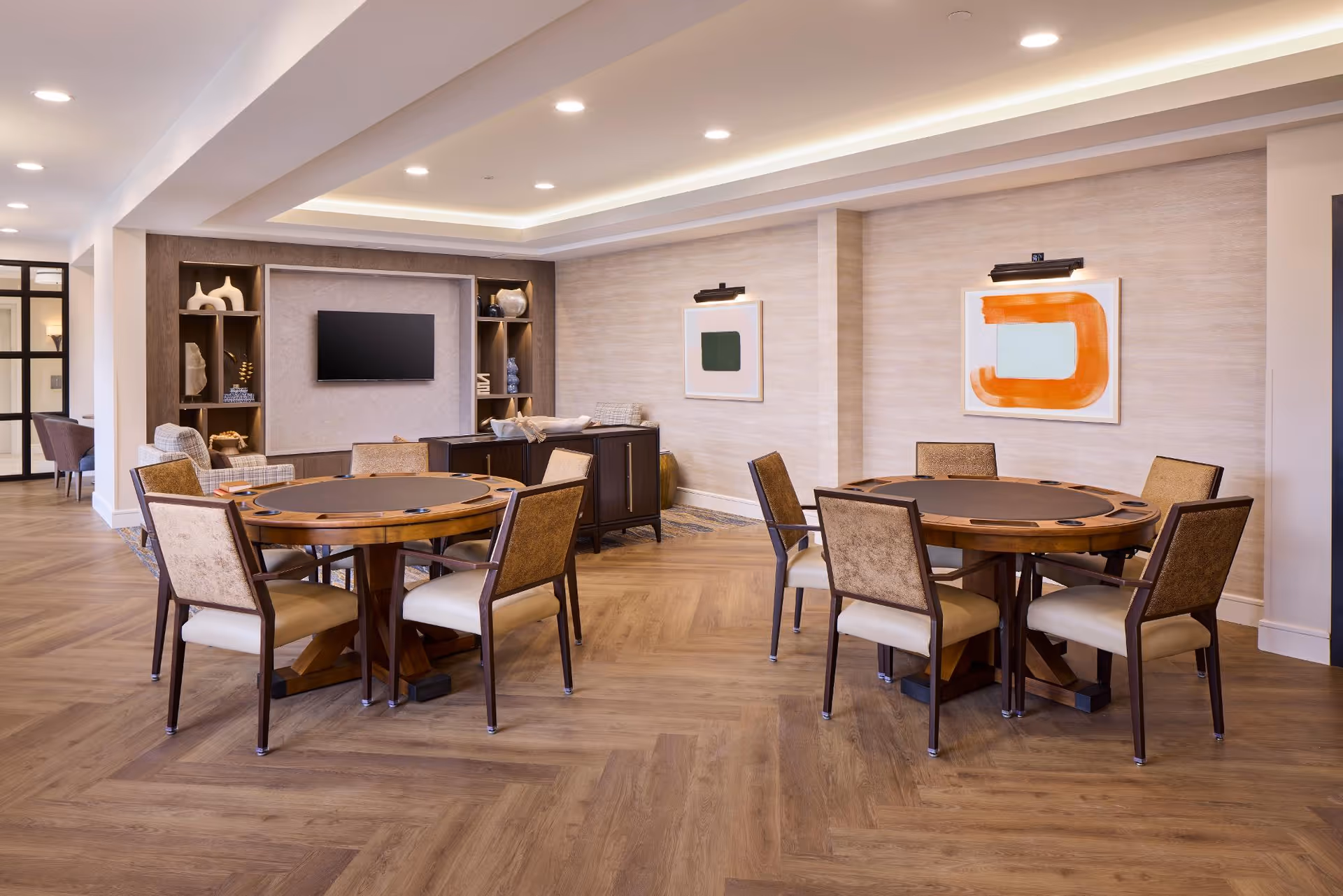 A spacious, well-lit common area with two round wooden tables each surrounded by six cushioned chairs. The room features wood flooring, modern recessed ceiling lights, and neutral-colored walls adorned with abstract artwork. A built-in shelving unit with decorative items and a wall-mounted TV is visible in the background.