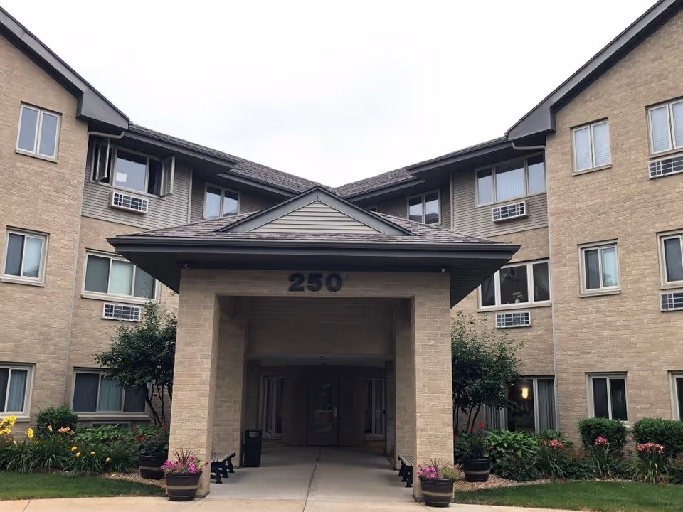 Front exterior view of a three-story senior living facility building with beige brick walls, multiple windows with air conditioning units, a covered entrance with the number 250 above it, and landscaped greenery including flowers and bushes on either side of the entrance.