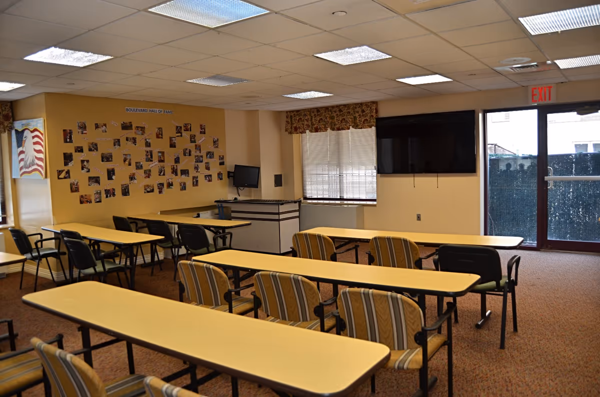 A room with several long tables and chairs arranged in rows facing a wall-mounted TV. The walls are beige with a bulletin board labeled 'Boulevard Hall of Fame' displaying many photos. There is a window with blinds and floral valance, and a glass door with an exit sign above it.