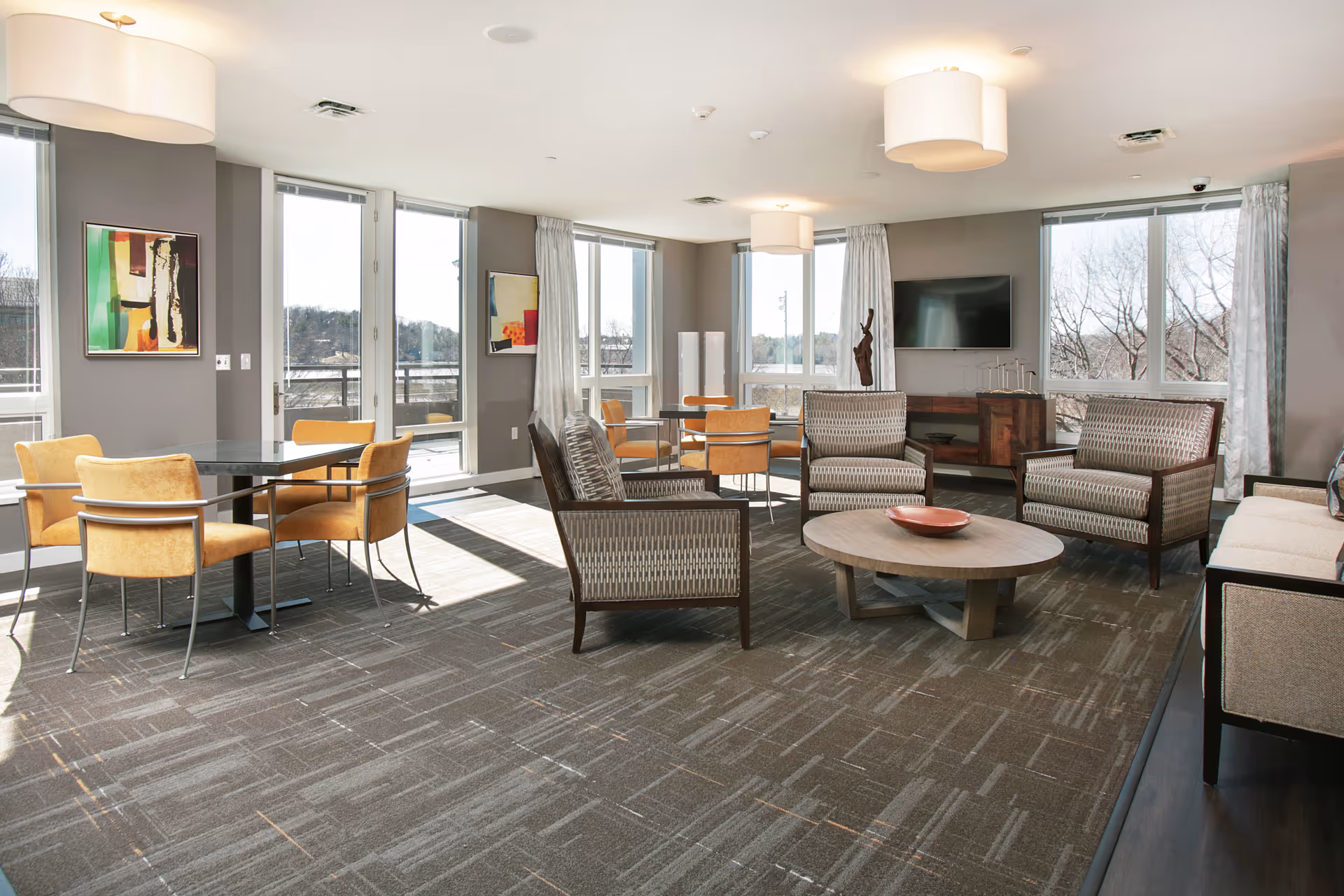 A bright and spacious common area with large windows letting in natural light. The room features several seating arrangements including patterned armchairs around a round wooden coffee table, a beige sofa, and tables with mustard yellow chairs. Modern ceiling lights hang from the white ceiling, and abstract art pieces adorn the gray walls. A flat-screen TV is mounted on one wall above a wooden console, and outside the windows, trees and a clear sky are visible.