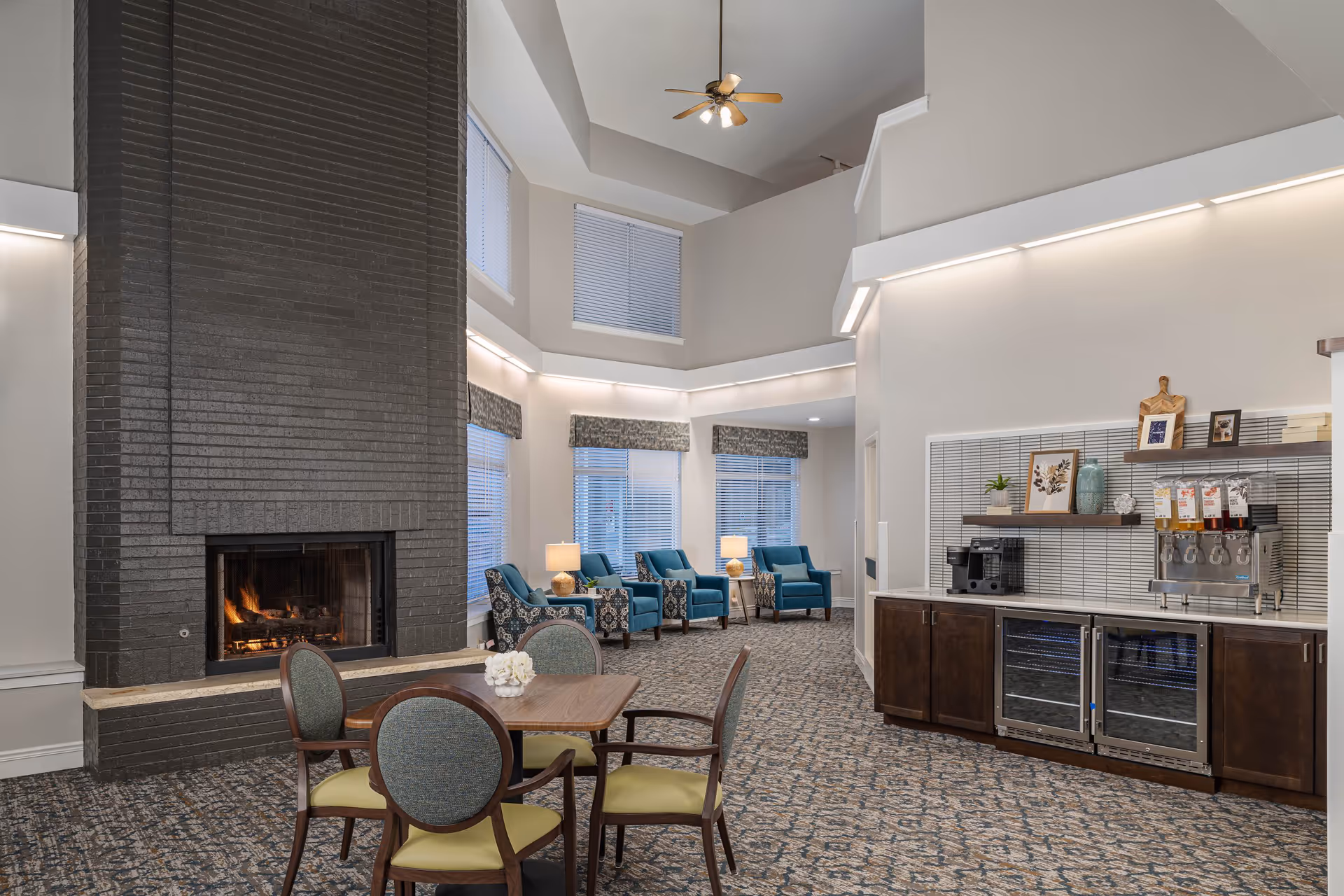 A cozy senior living common area with a tall black brick fireplace, a wooden table with four chairs, and a seating area with blue armchairs near large windows. On the right side, there is a refreshment station with a coffee machine, beverage dispensers, and two glass-front refrigerators beneath a shelf with decorative items.