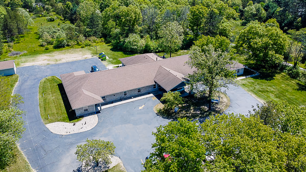 Aerial view of a single-story senior living facility building surrounded by trees and greenery, with a paved driveway and parking area.