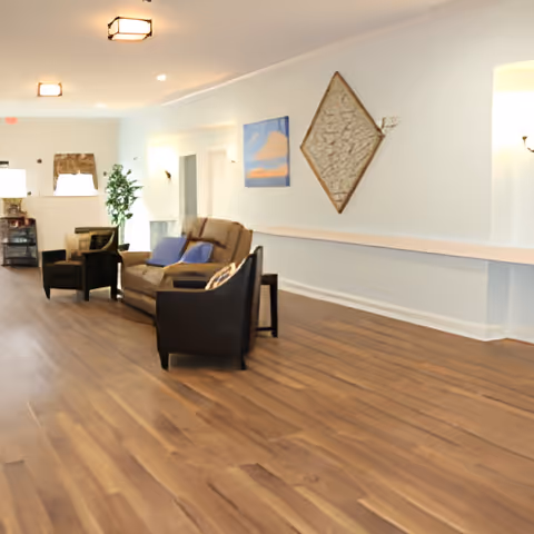 A spacious senior living facility common area with wooden flooring, a brown sofa with blue cushions, two black armchairs, wall decorations including a diamond-shaped piece and a painting, and a potted plant near the window.
