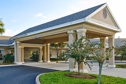 Entrance of Solaris HealthCare Windermere facility showing a covered driveway with columns, a landscaped area with small trees and shrubs, and a clear sky above.